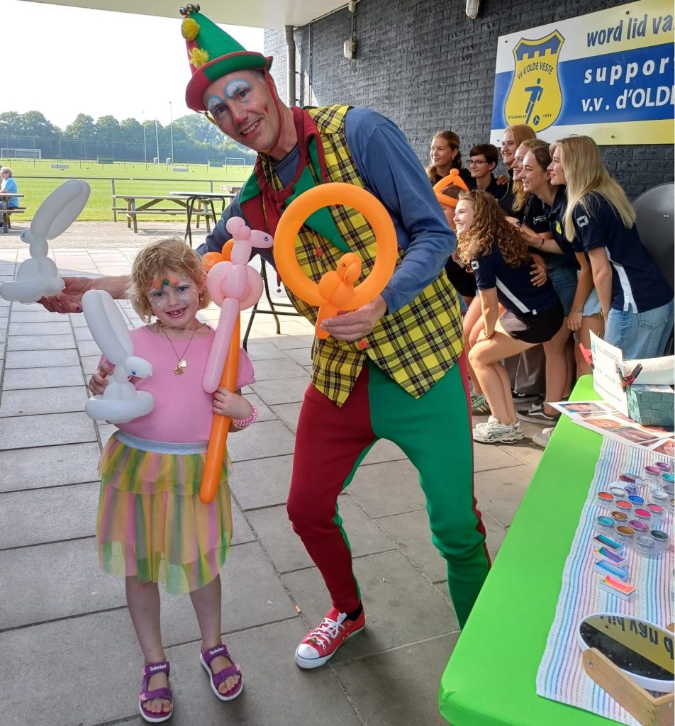 Ballonnenclown in harlekijncostuum geeft geschminkt meisje een ballon papagaai in Steenwijkerland